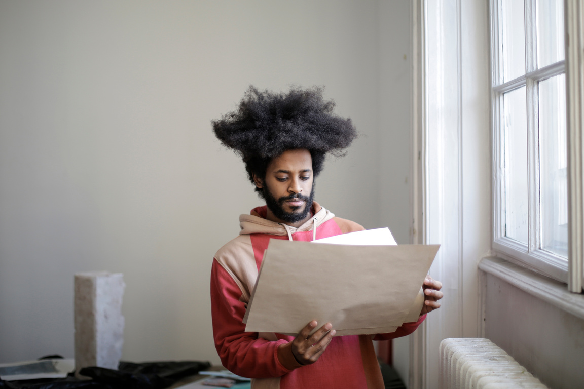 Serious young ethnic male worker holding papers near window in workshop